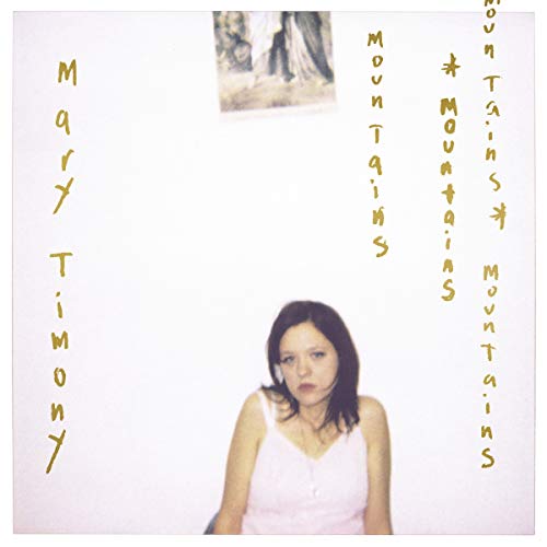 A woman in a white camisole stands against a white wall with gold text.