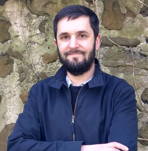 Man with dark hair and beard, arms crossed, against a stone wall.