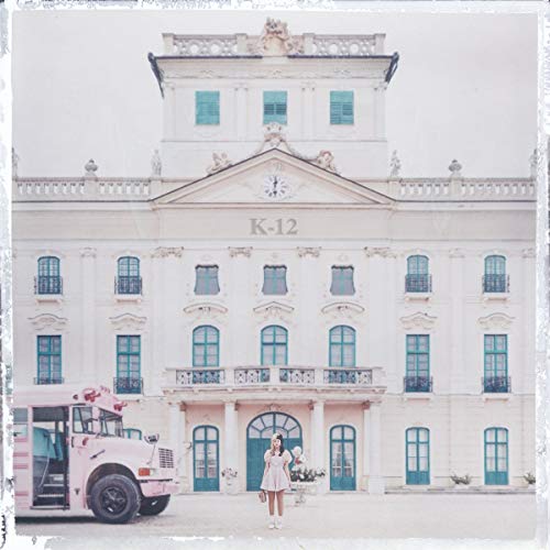 The image depicts a large, white building with a clock on the front and a pink bus parked in front of it. A woman stands in the foreground, wearing a white dress. The building has multiple windows and a door, and the overall atmosphere is one of elegance and sophistication. The image is likely an album cover for Melanie Martinez's "K-12".