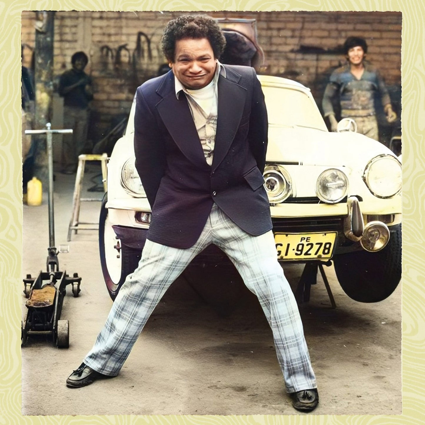 A man in a blazer and plaid pants poses in front of a car.