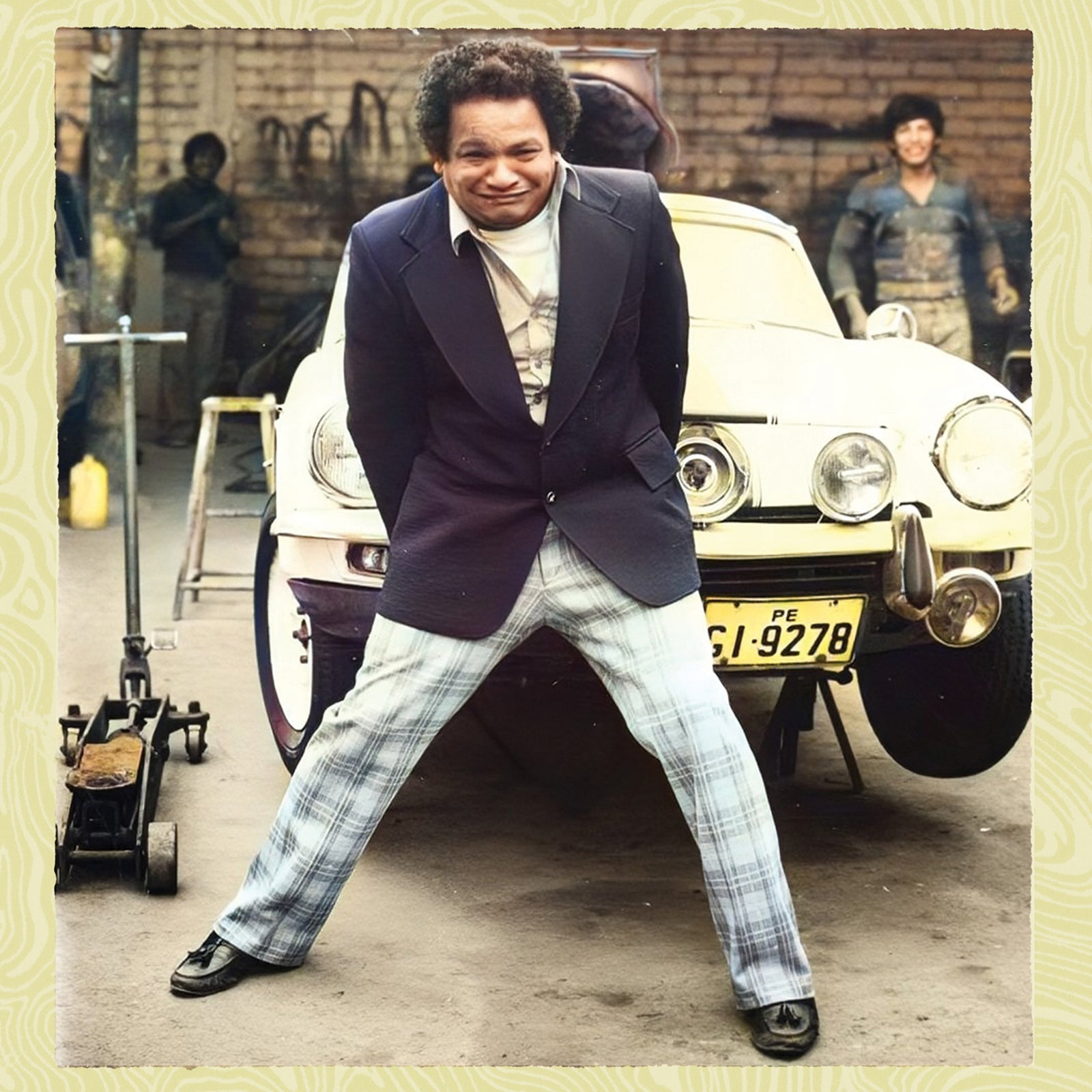 A man in a blazer and plaid pants poses in front of a car.