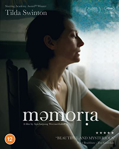 Woman with short dark hair sitting in chair looking right, white shirt, window behind.