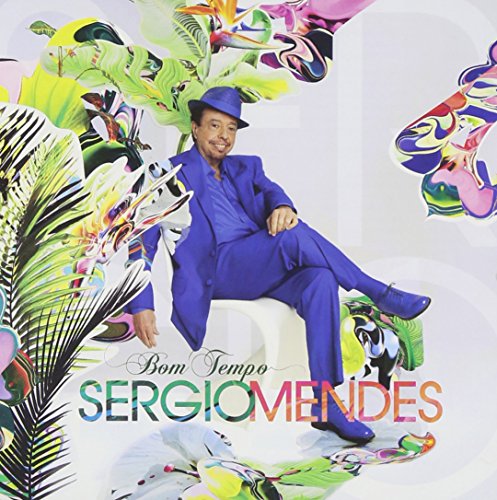 Sergio Mendes in a blue suit sits amidst vibrant tropical foliage.