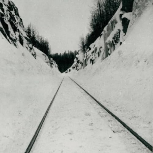 Black and white photo of snowy railroad tracks between tall snowbanks.