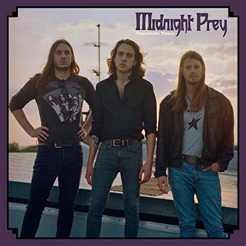 Three men with long hair standing outdoors against a sunset sky.
