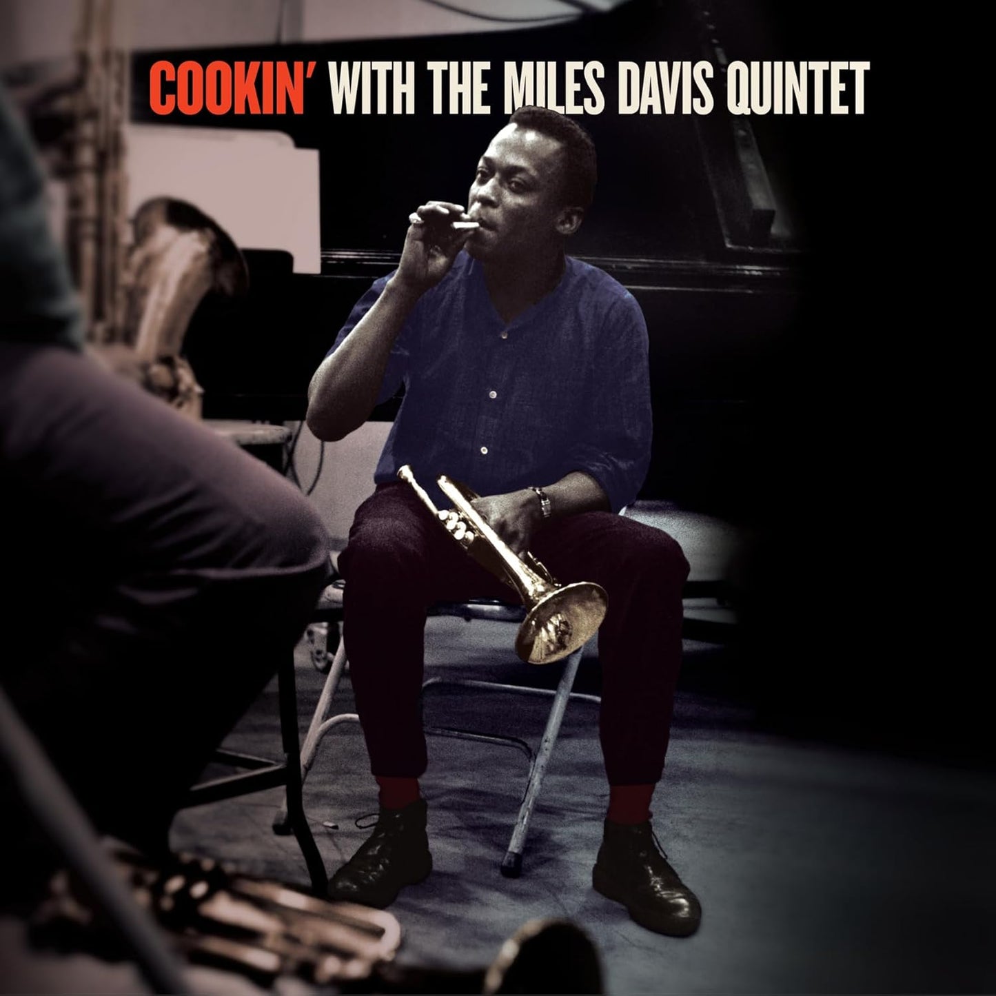 Miles Davis seated, holding a trumpet, with band members in the background.