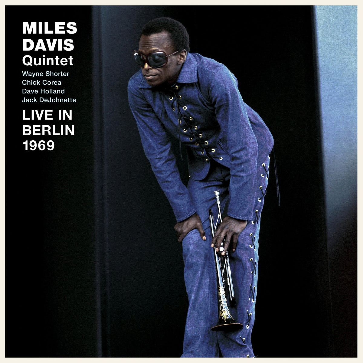 Miles Davis in a blue denim outfit holding a trumpet against a black background.