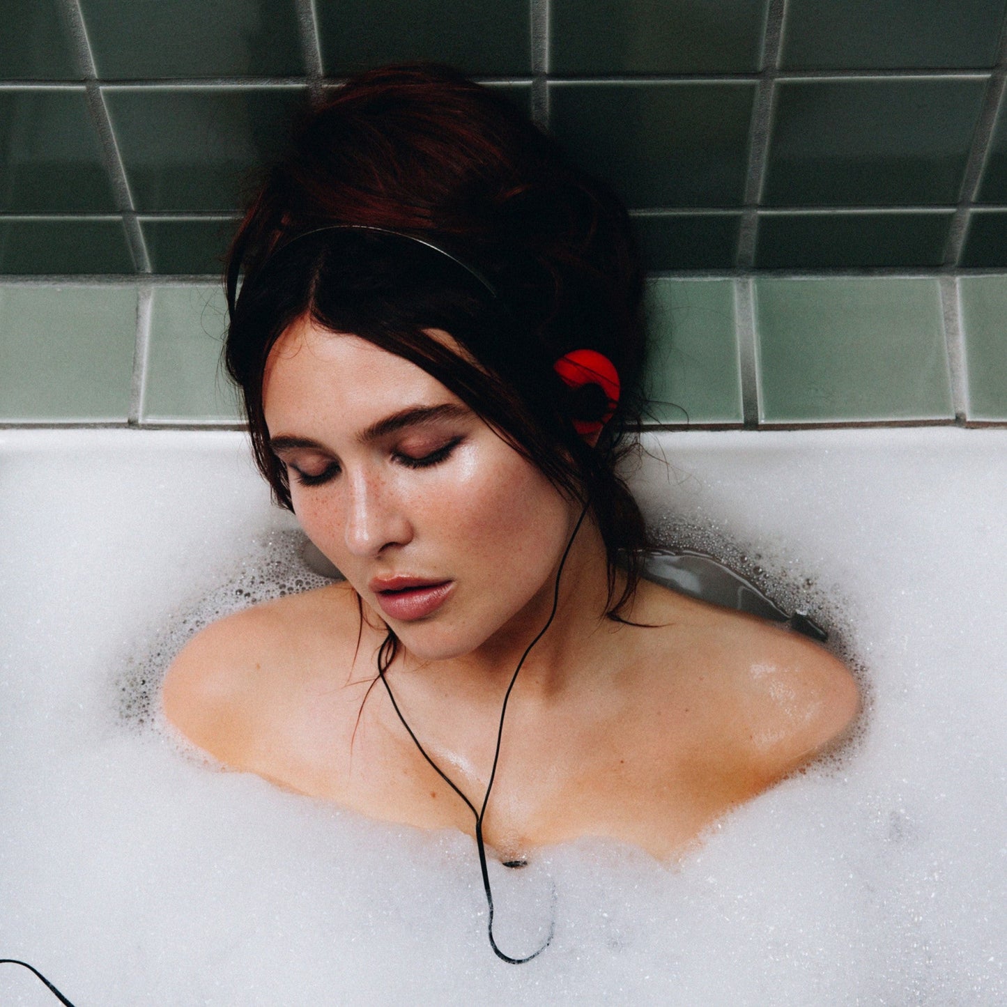 Woman with red headphones lies in a bubbly bathtub, eyes closed.