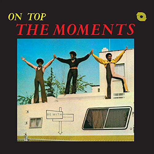 Three men in matching outfits stand with arms raised on top of a camper.