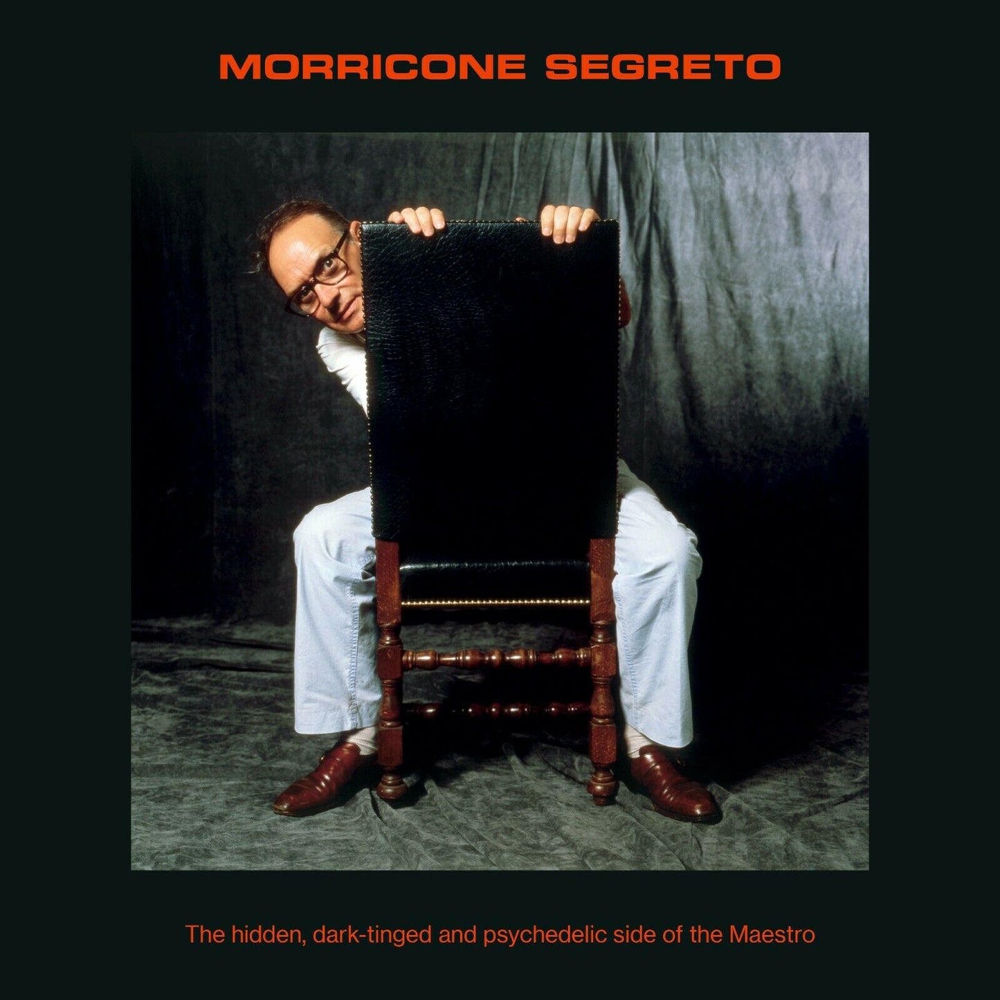 "Album cover with a man hiding behind a chair, dark background, and orange text."