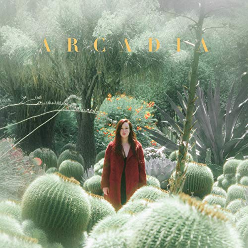 Woman in red coat stands among cacti and lush greenery.