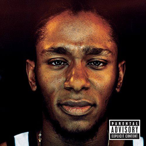 Close-up portrait of Mos Def, with a dark background and warning label.