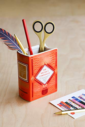 A book-shaped orange ceramic vase holding a feather, a red pencil, and scissors.