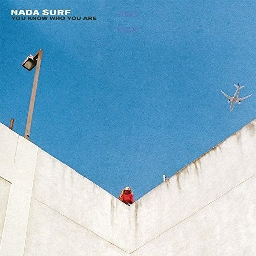 Album cover: person on a rooftop with a plane in a blue sky.
