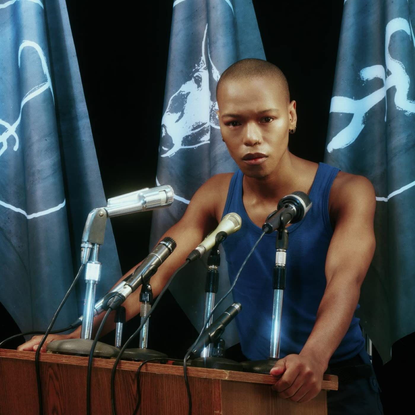 Man with shaved head stands behind podium with multiple microphones, blue flags behind.