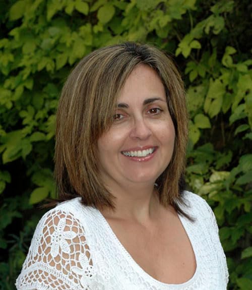 ASSPC Smiling woman with brown hair wearing a white lace top against green foliage.