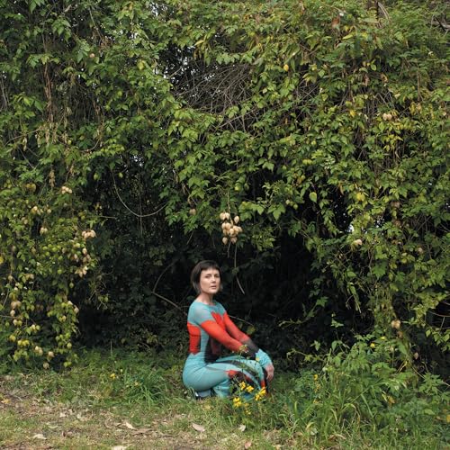 Woman in patterned bodysuit sits amongst lush green foliage.