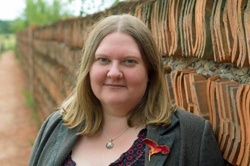 A woman with shoulder-length blonde hair stands in front of a weathered brick wall.
