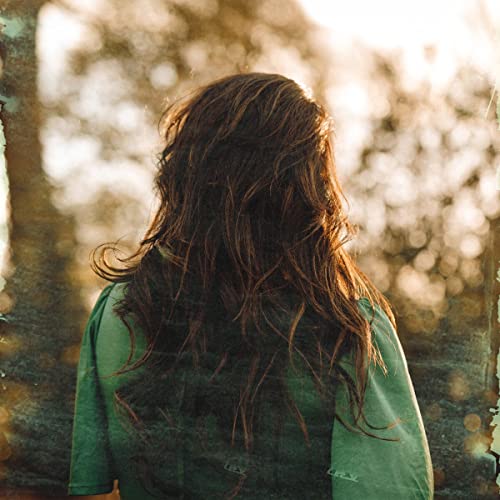 A woman with long brown hair, seen from behind, wearing a green shirt outdoors.