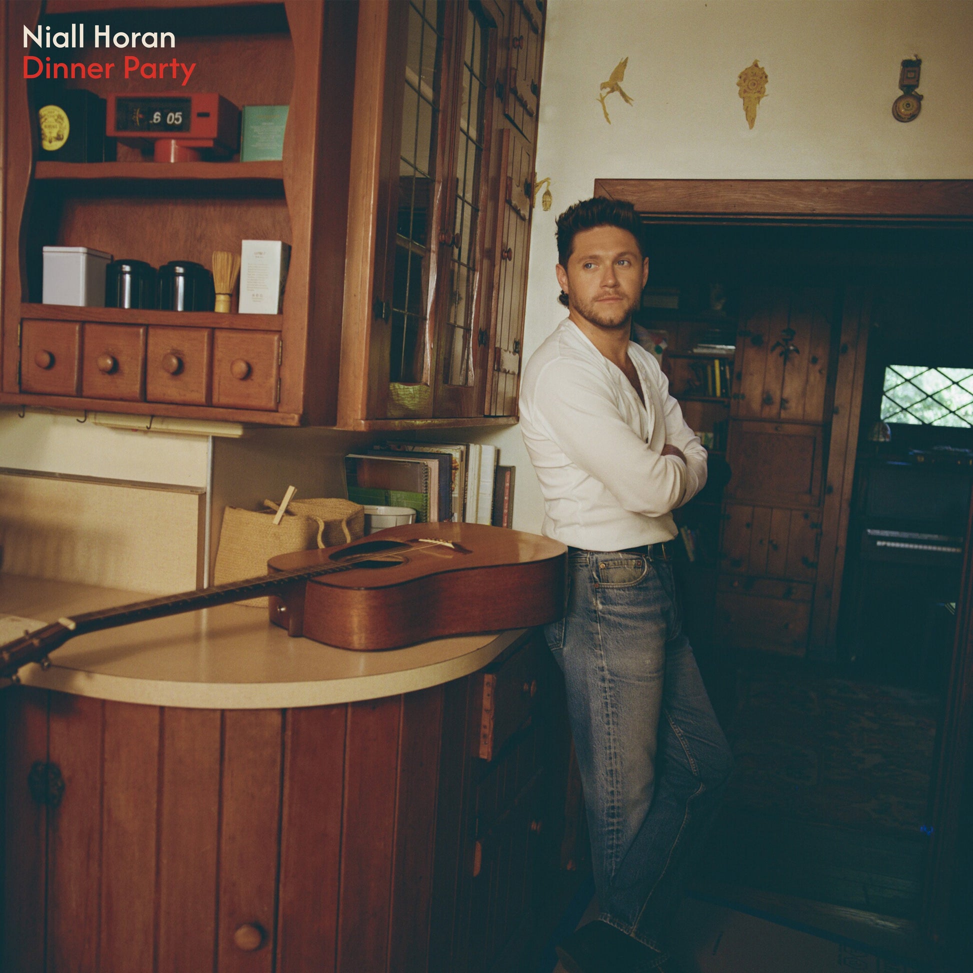 Niall Horan poses in a kitchen with wooden cabinets and a guitar.