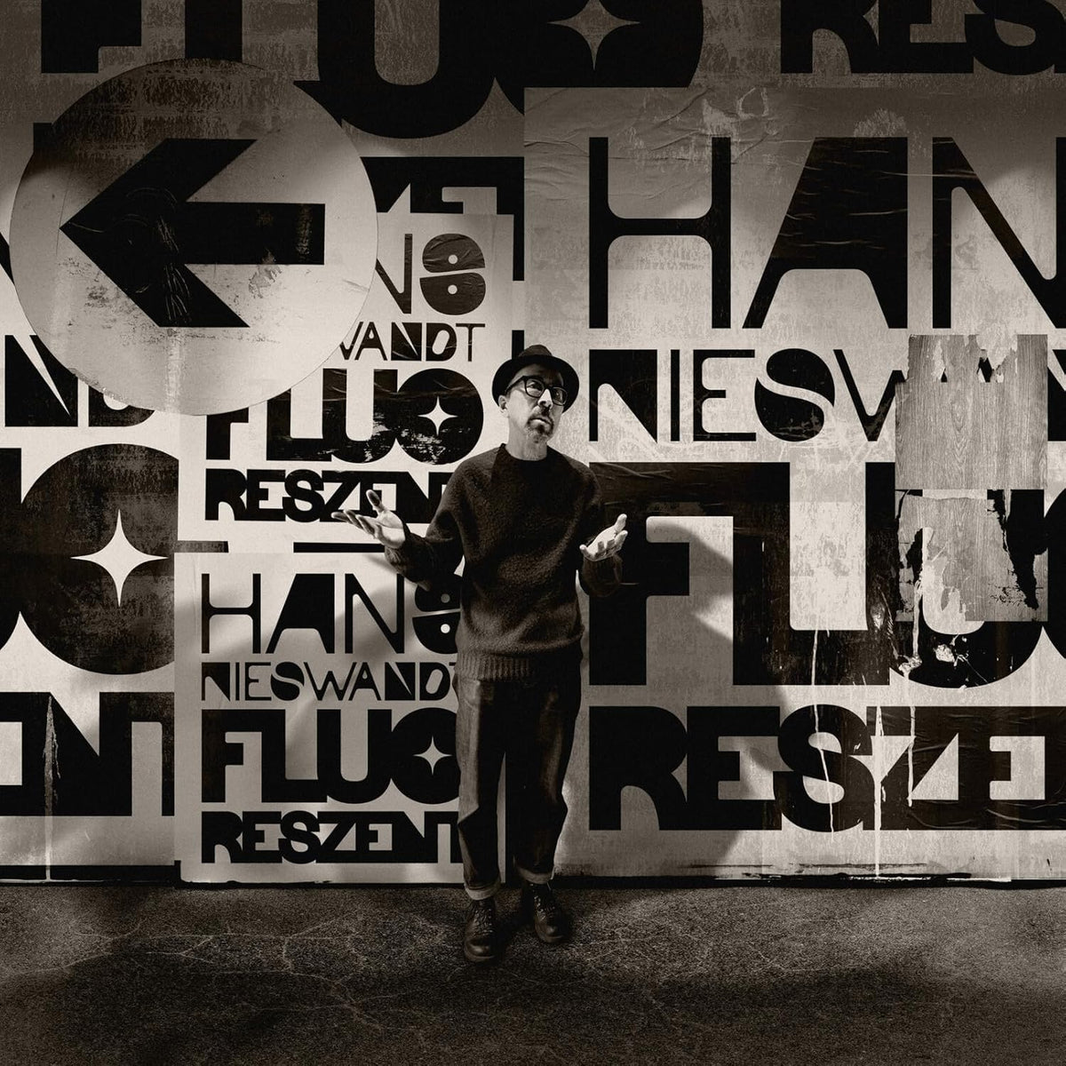 A man in a hat and sweater stands before a wall of bold typography.
