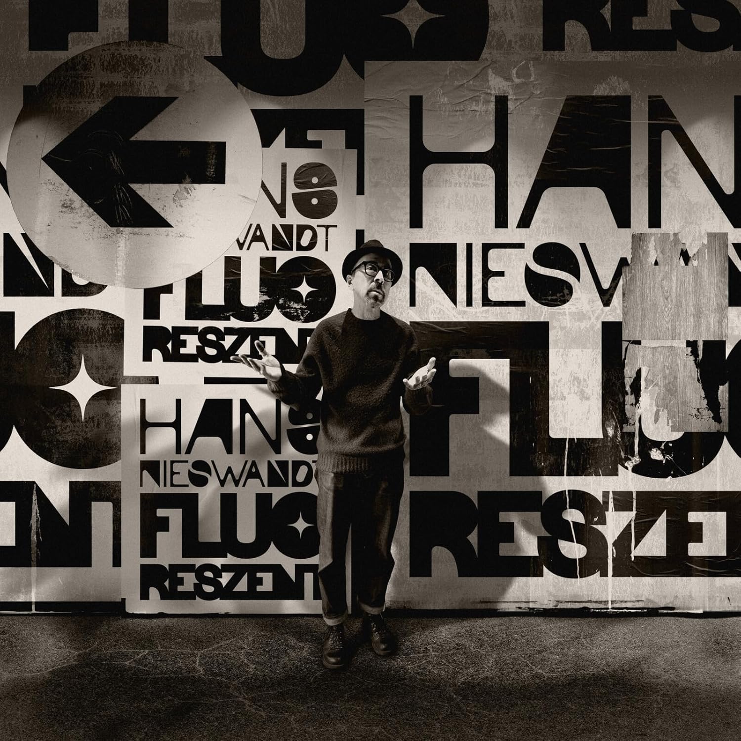 A man in a hat and sweater stands before a wall of bold typography.