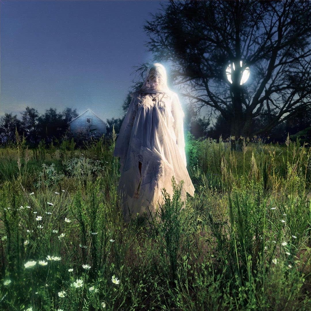Woman in white gown stands in a field under a tree.