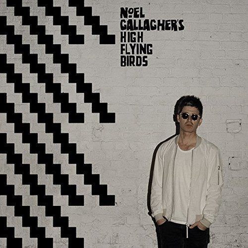 Noel Gallagher in white jacket and sunglasses against a white brick wall with black stripes.
