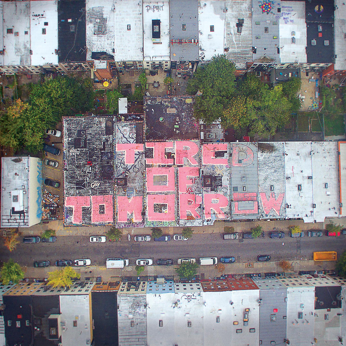 Aerial view of rooftops with pink graffiti reading "TIRED OF TOMORROW".