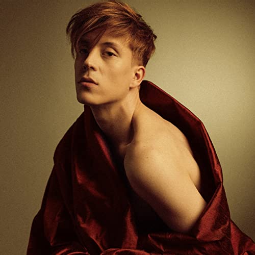 Young man with reddish hair, partially draped in red fabric.
