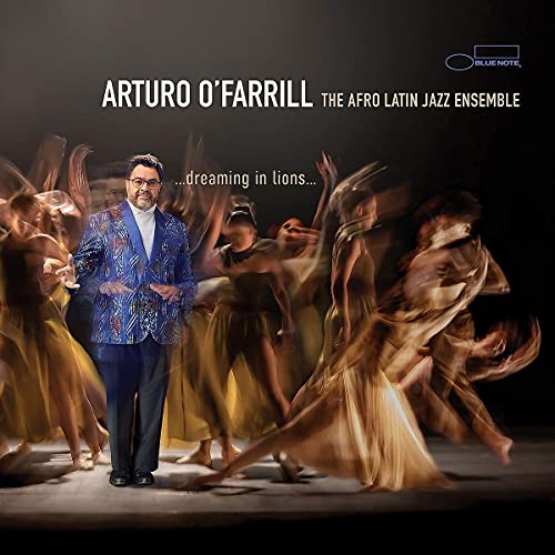 Arturo O'Farrill in a blue patterned blazer standing before blurred dancers in gold dresses.
