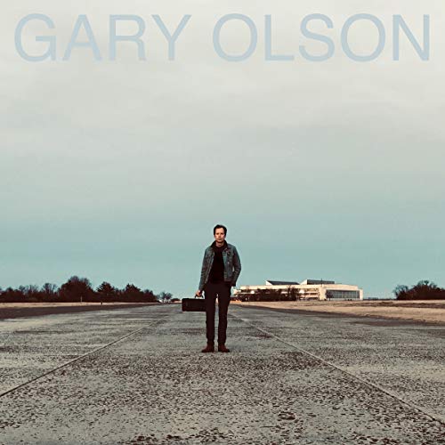 "Album cover: man standing on a runway with a suitcase, building in the background."