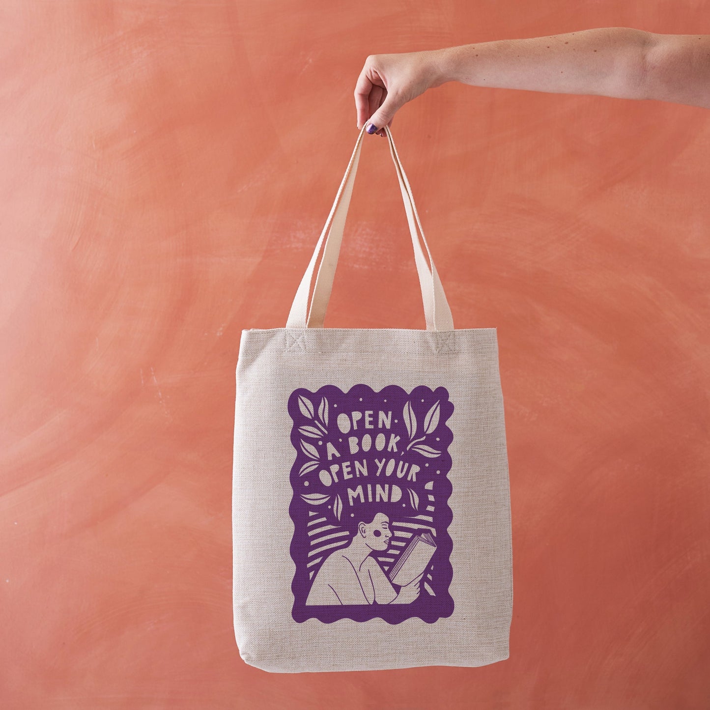 Beige tote bag with purple illustration of a person reading a book.