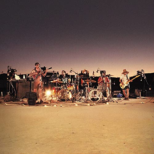 Band performing outdoors at dusk, with drums, guitars, and stage lighting.