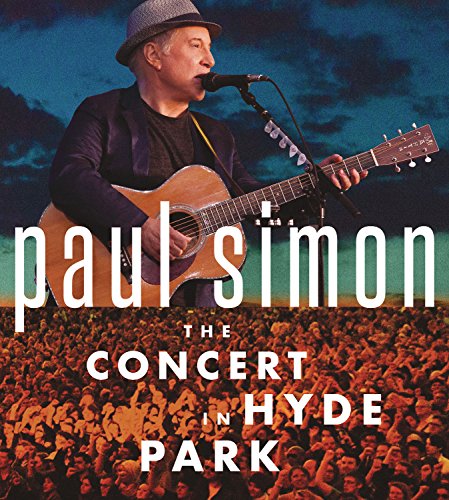 Paul Simon wearing a hat and playing acoustic guitar before a large concert crowd.