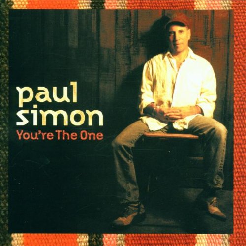 Paul Simon album cover: man seated on stool, dark background, orange border.