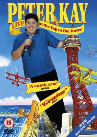 Peter Kay holding a microphone in front of the Blackpool Tower with biplanes flying.