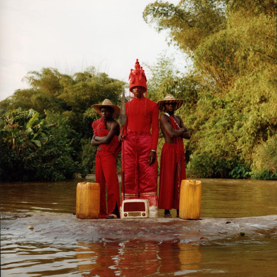 Three figures in red stand on a raft in a river landscape.
