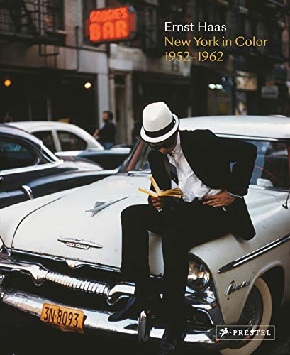 A man in a fedora sits on a white vintage car reading a book.