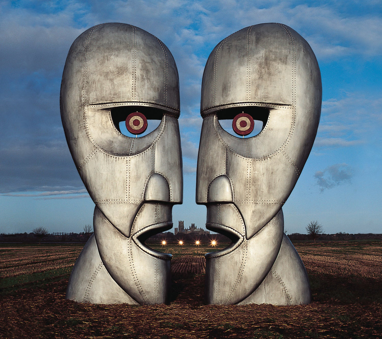 Two large metal heads with glowing eyes face each other in a field.