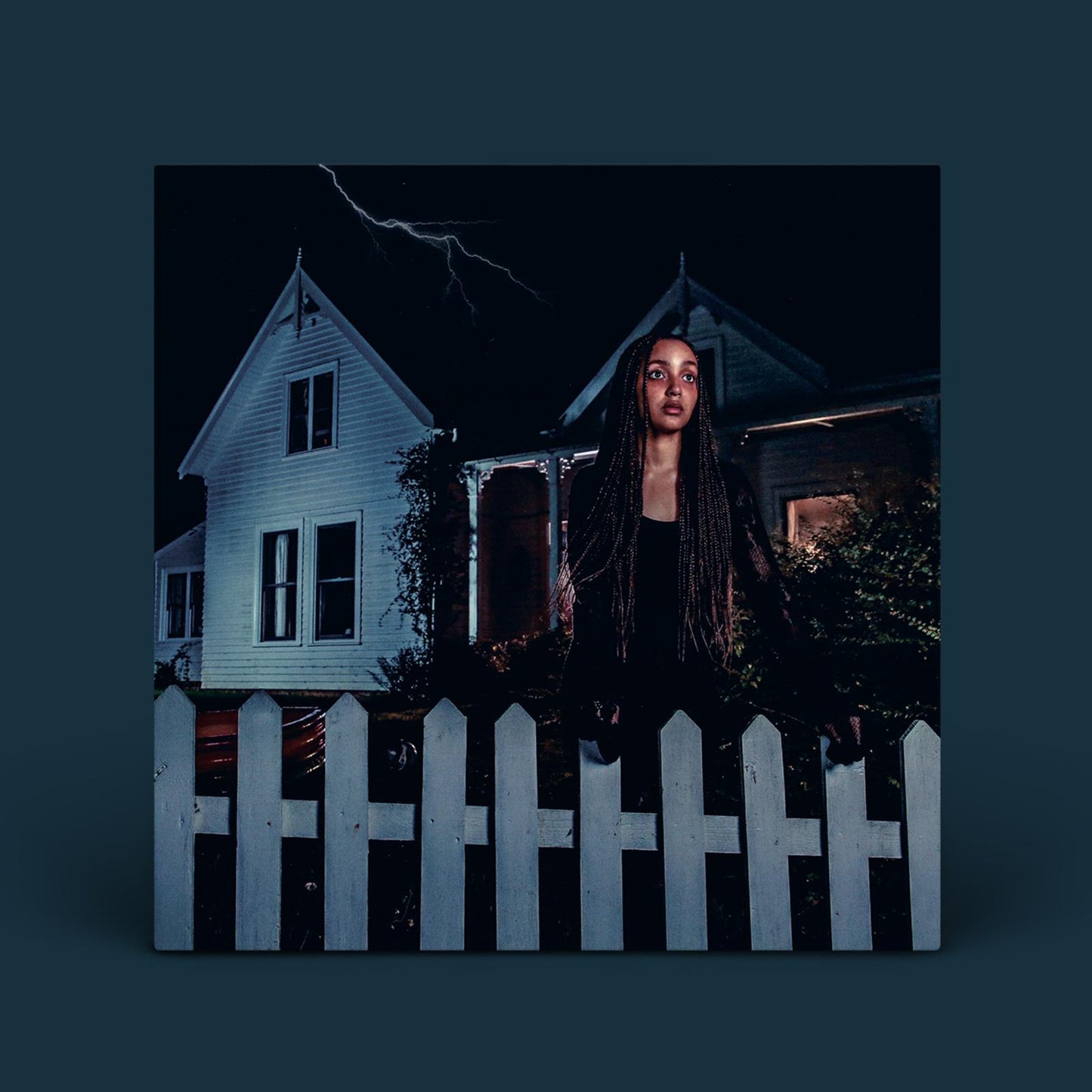 PinkPantheress stands behind a white picket fence in front of a house at night.