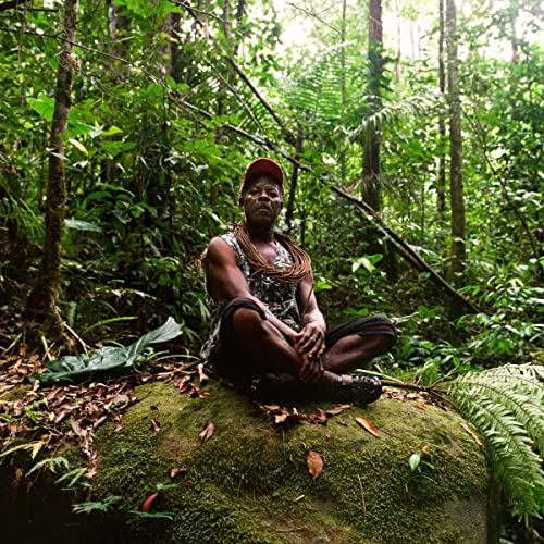 Man with red cap seated on a rock in lush forest.