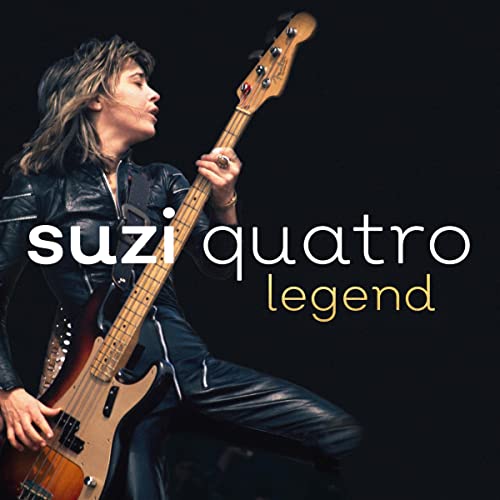 Suzi Quatro in a black leather outfit playing a bass guitar against black background.