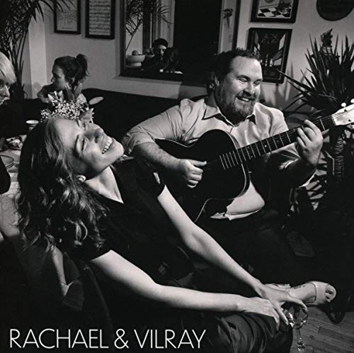 Black-and-white image of a man playing guitar beside a reclining woman, indoors with others.