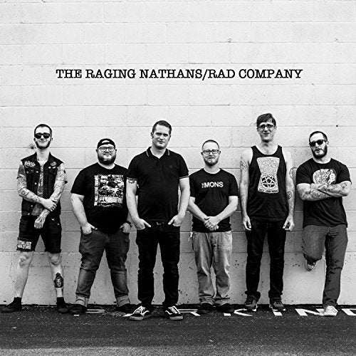 Six men stand in a row against a white brick wall in this photograph.