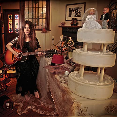 Woman with guitar, dolls, and a tiered cake in a dimly lit room.