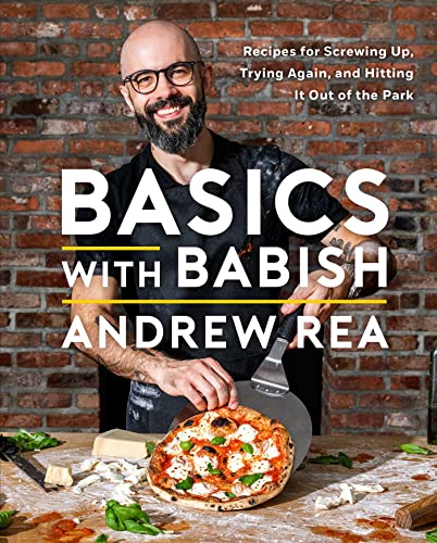 Andrew Rea smiling in a black apron, holding a pizza peel over a pizza.