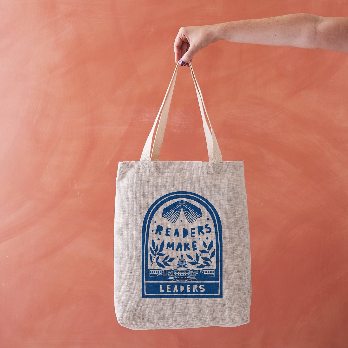 A hand holds a beige canvas tote bag with a blue graphic of books.