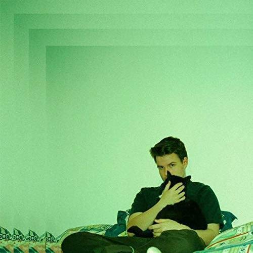 A man sits on a patterned couch, wearing a black shirt, against a green background.