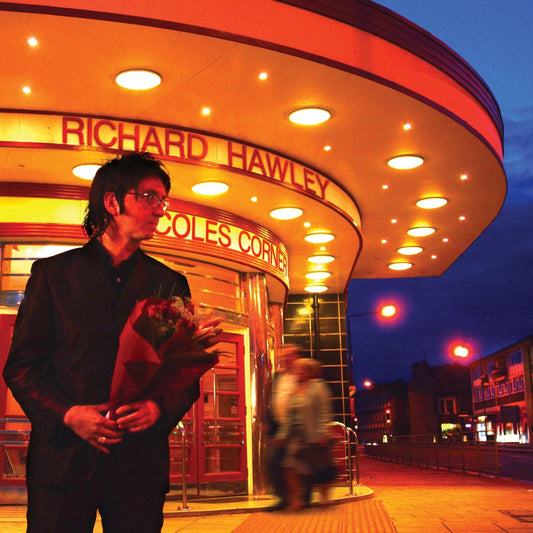 Richard Hawley stands before a brightly lit cinema marquee, holding flowers.